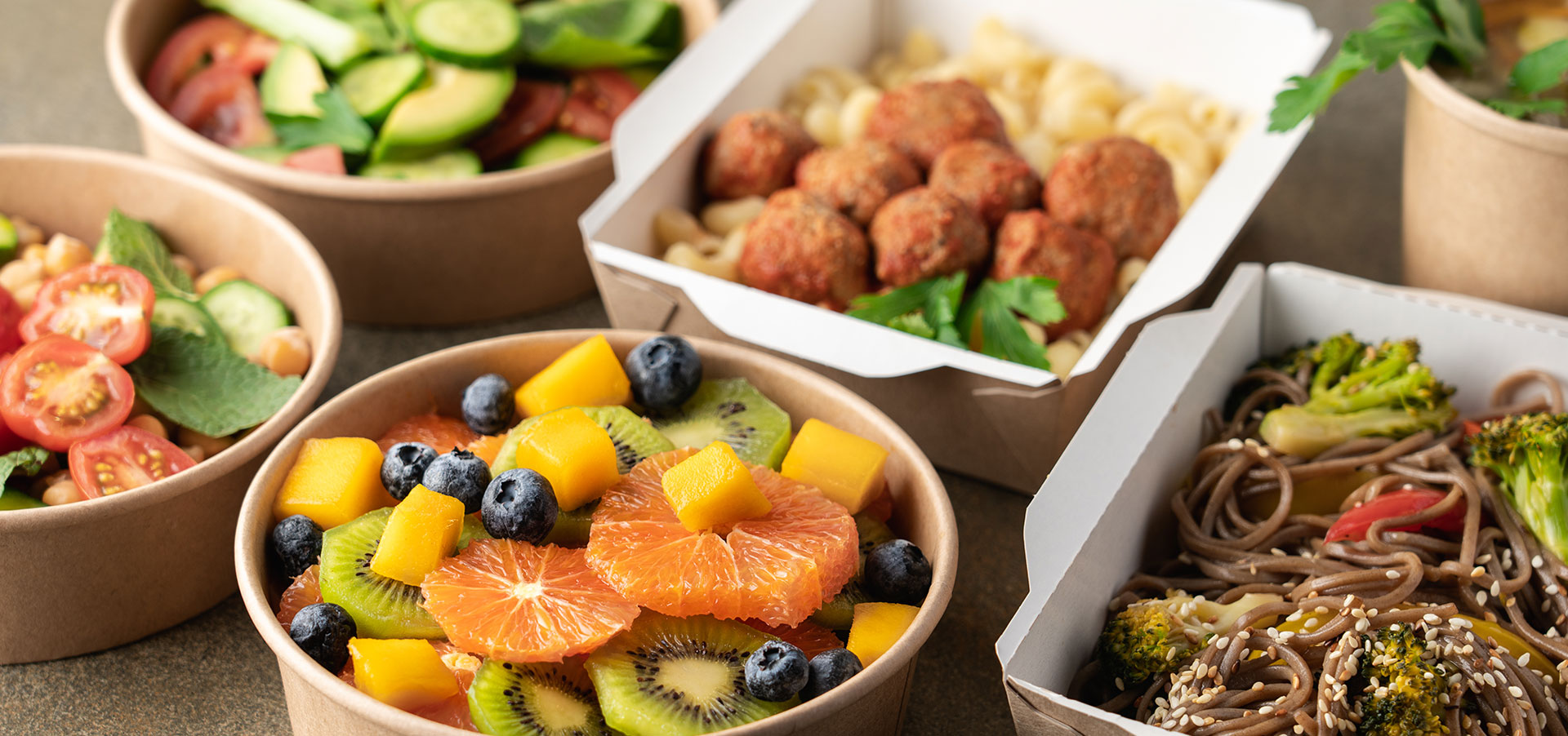 Recycled paper bowls and food containers hold green salads with tomatoes, cucumber, and avocado, a fruit salad with kiwi, mango, blueberries, and tangerine, a broccoli and noodle dish with sesame seeds, a meatball and macaroni dish with parsley