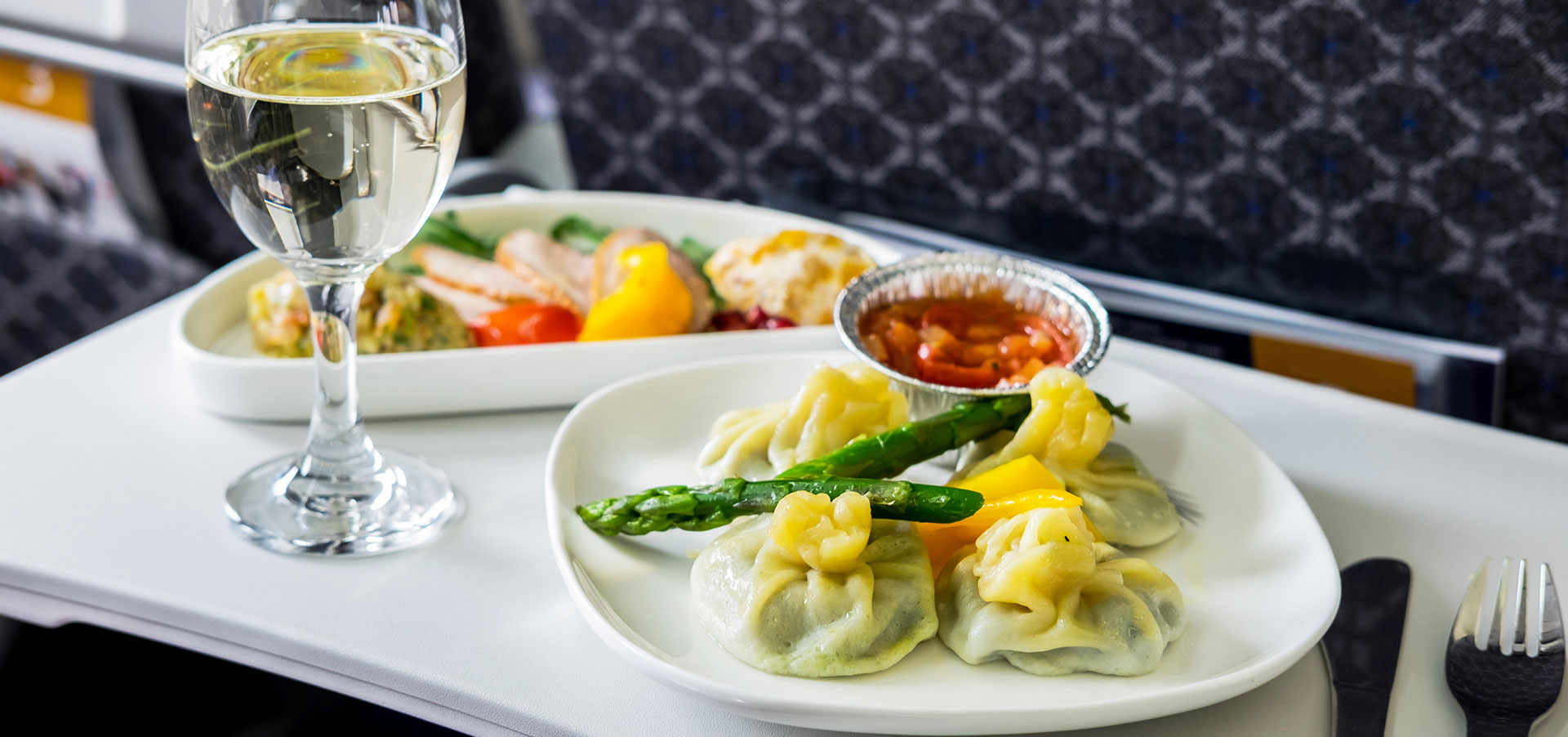Airplane tray holds an exquisite meal of ravioli with asparagus, chicken with squash and a rice pilaf, with a stemmed glass goblet of water and silverware